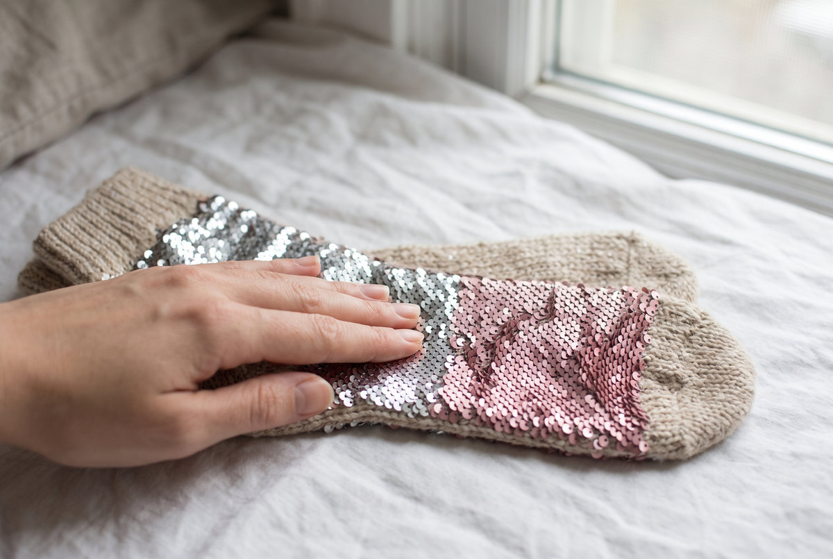 Main qui brosse les paillettes réversibles d'une chaussette posée sur un lit, les paillettes changeant de couleur sous le doigt