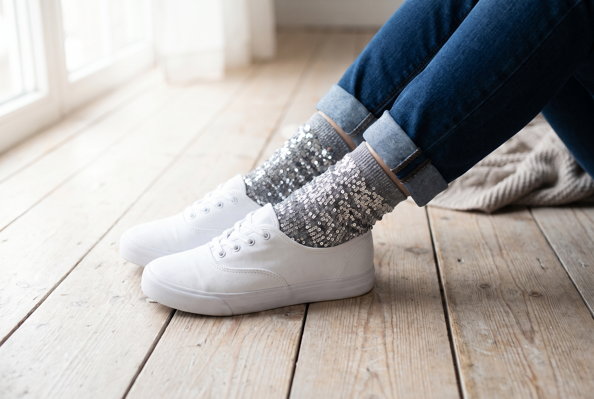 Pieds croisés portant des chaussettes à paillettes grises aux reflets argentés, avec un jean retroussé et des baskets blanches sur un parquet clair