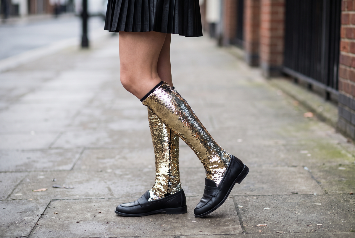 Jambes vues de côté portant des chaussettes hautes à paillettes dorées sous une jupe plissée noire, avec des mocassins noirs